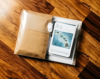 May include: A clear plastic envelope with a brown paper envelope inside. The brown envelope has a white button closure. The clear envelope contains a white card with the letter "A" and the word "archipelago" printed on it. The card has a watercolor illustration of an archipelago.