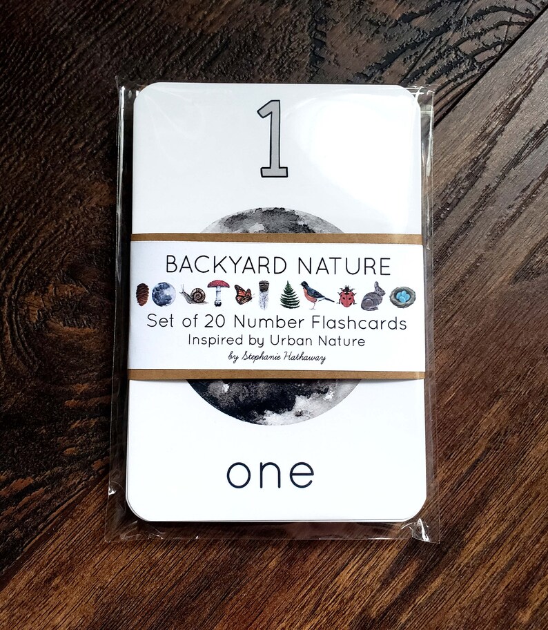 May include: A white flashcard with a black and white illustration of a moon. The number "1" is printed in black at the top of the card. The text "BACKYARD NATURE" is printed in brown on a tan background. The text "Set of 20 Number Flashcards Inspired by Urban Nature by Stephanie Hathaway" is printed in black on a tan background. The word "one" is printed in black at the bottom of the card.