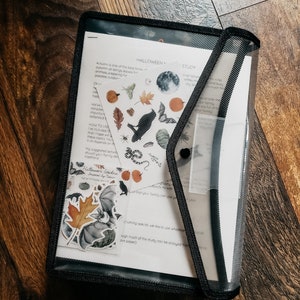 May include: A clear plastic document holder with a black border, containing printed pages and stickers with a Halloween theme. The pages feature text and illustrations of autumn leaves, bats, and pumpkins. The holder is on a wooden surface.