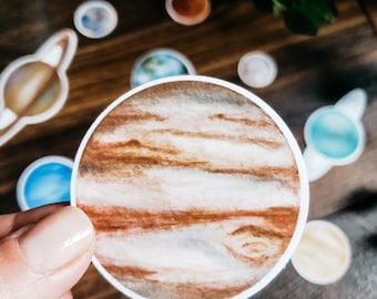 May include: A hand holds a round sticker depicting Jupiter, with brown and white bands. Other planet stickers are scattered on a wooden surface, including Saturn, Earth, and Mars. The stickers are watercolor style.