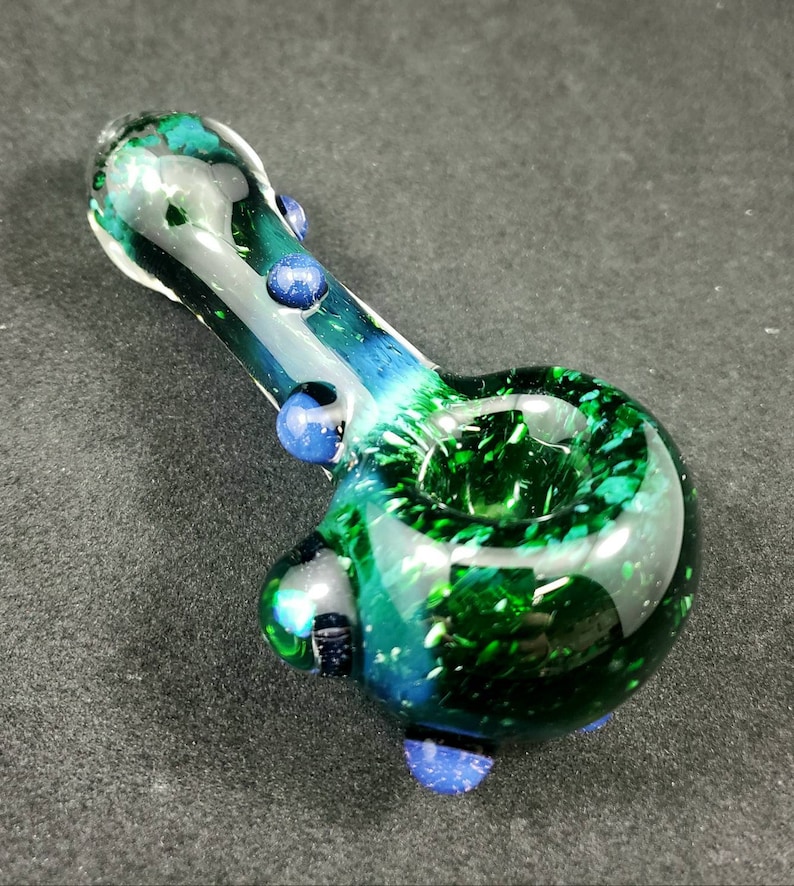 May include: A green glass pipe with blue accents. The pipe has a round bowl and a long, thin stem. The bowl is decorated with green and blue speckles.