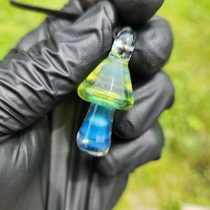 May include: A glass mushroom pendant with a yellow and green cap and a blue and white stem. The pendant is hanging from a black cord.