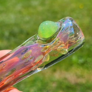 Puede incluir: Una pipa de vidrio con una esfera verde iridiscente en el extremo. La pipa está hecha de vidrio transparente con remolinos rosas y naranjas.