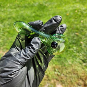 Może przedstawiać: Zielona szklana fajka z zaokrągloną cybuchą i długą, smukłą rurką. Fajka ma ciemną, okrągłą cechę w cybuchu. Fajka jest trzymana w rękawicy na tle zielonej trawy.