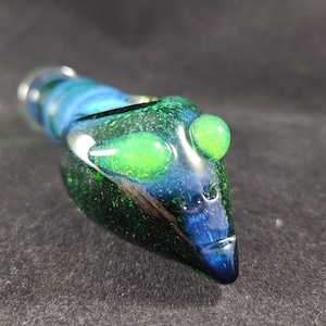 May include: A green and blue glass pipe with a unique alien-like design. The pipe has three green eyes and a textured surface.