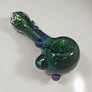 May include: A green glass pipe with white and blue speckles. The pipe has a round bowl and a long, thin stem. There are three small blue and purple glass accents on the pipe.