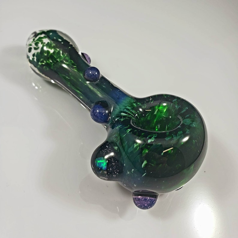 May include: A green glass pipe with white and blue speckles. The pipe has a round bowl and a long, thin stem. There are three small blue and purple glass accents on the pipe.