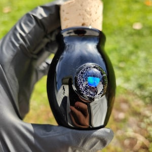May include: A black glass bottle with a cork stopper. The bottle has a round, clear glass bubble with a blue and white design. The bottle is held by a person wearing black gloves. The background is blurred green grass.