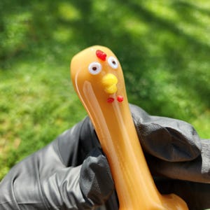 May include: A novelty glass pipe in the shape of a chicken. The pipe is primarily amber-colored with white, yellow, and red accents. The chicken's face features white eyes, a yellow beak, and red waddles. The pipe is held by a gloved hand.