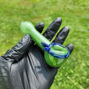 May include: A green glass pipe with a rounded bowl and a long stem. The bowl is accented with a dark blue band and a small blue decorative element. The pipe is held in a black gloved hand.