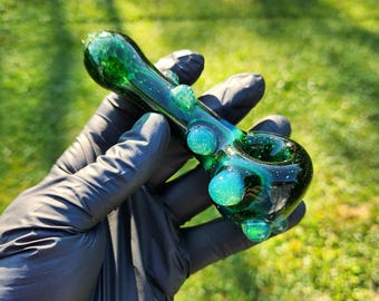 May include: A green and blue glass pipe with a speckled design. The pipe has a bowl, a stem, and several rounded embellishments. The glass is transparent, allowing light to pass through. The pipe is held in a gloved hand against a green background.