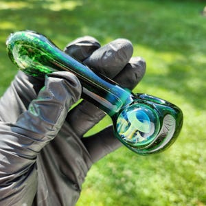 May include: A hand-blown glass pipe with a green and blue color scheme. The pipe features a rounded bowl with a decorative design and a long, curved stem. The glass has a marbled appearance with swirls of color.