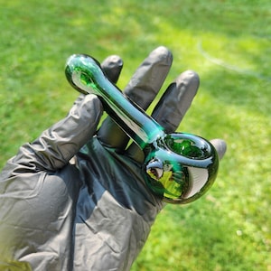 May include: A green glass pipe with a rounded bowl and a long stem. The pipe is held in a gloved hand against a backdrop of green grass. The glass has a glossy finish.