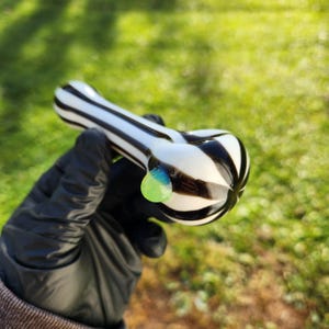 May include: A black and white striped glass pipe with a green and blue accent. The pipe is held in a gloved hand.
