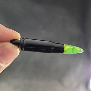 May include: A black bullet casing with a green glitter-filled tip. The casing is shiny and has a smooth surface.
