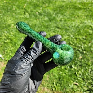 May include: A green glass pipe with a speckled pattern and a rounded bowl. The pipe has a small, round, light green accent on the bowl. The pipe is held in a gloved hand against a background of green grass.