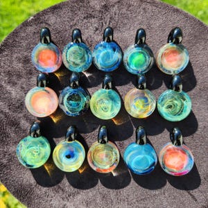 May include: A selection of colourful glass pendants with black tops. Each pendant has a unique swirling design in shades of blue, green, orange, and yellow. The pendants are displayed on a dark brown surface.