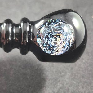 Puede incluir: Pipa de vidrio negro con un diseño de galaxia. La pipa presenta un cuenco redondeado con un patrón arremolinado de azules, blancos y dorados. El tallo tiene forma cilíndrica y acanalada.