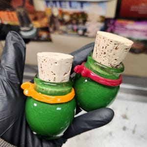 Puede incluir: Dos botellas de vidrio verde con tapones de corcho. Una botella tiene una banda naranja, la otra una banda roja. Las botellas son sostenidas por una mano enguantada. Las botellas son pequeñas y redondas.