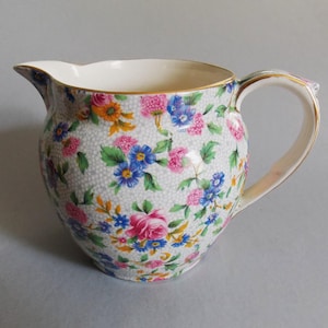 May include: A ceramic creamer pitcher featuring a white background with a fine grey honeycomb pattern and a colorful floral print of pink roses, blue forget-me-nots, and orange blossoms. The rim and handle are accented with a thin gold trim.