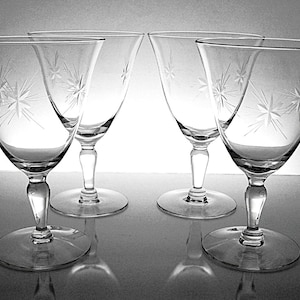 May include: Four clear stemmed wine glasses with starburst designs etched into the bowls. The glasses have a classic shape with a rounded bowl and a slender stem. The glasses are arranged in a symmetrical pattern.