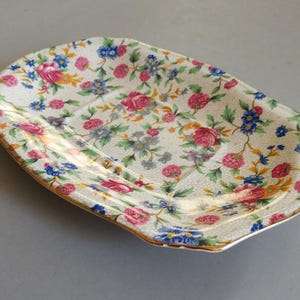 May include: An oval ceramic dish featuring a cream-colored background with a dense pattern of pink roses, blue forget-me-nots, and green foliage. The dish has a subtle crackle glaze finish and a thin gold-toned trim along the scalloped edges.