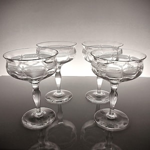 May include: A set of four clear glass coupe glasses, each featuring a delicate etched pattern of interconnected loops and swags around the bowl. The glasses have a wide, shallow bowl shape, a slender stem with a subtle curve, and a circular base.