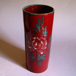 May include: A tall, cylindrical red ceramic vase with a floral design. The vase features a large, white and red flower with green leaves and accents. The rim has a green trim.