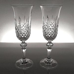 May include: A pair of clear crystal champagne flutes featuring a detailed diamond-cut pattern on the lower bowl, a faceted hexagonal stem node, and a smooth circular base. The upper portion of the bowl is plain and flares slightly outward.