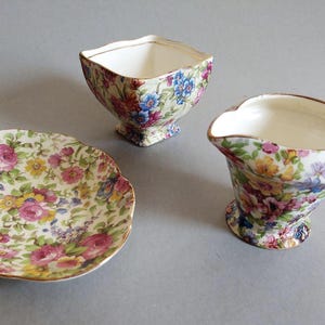 May include: A floral-patterned tea set featuring a small saucer, a square-shaped bowl, and a creamer with a handle. The set has a cream background with pink, blue, and yellow flowers and gold trim. The pieces are arranged on a gray surface.