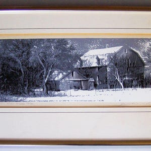 May include: Framed black and white print of a barn in a snowy landscape. The barn has a pitched roof covered in snow, and is surrounded by trees. The print is in a gold-colored frame.