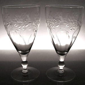 May include: Two clear, stemmed wine glasses with etched floral and geometric designs. The glasses have a rounded base, a bulbous stem, and a flared bowl. The etched design features leaves and geometric patterns. The glasses are displayed on a reflective surface.