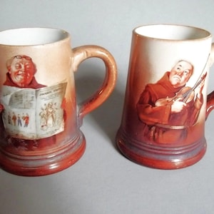 May include: Two ceramic mugs with a reddish-brown gradient finish. The left mug features a monk reading a newspaper, while the right mug depicts a monk playing a violin. Both mugs have a white interior, a sturdy handle, and a wide base design.