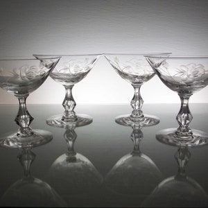 May include: Four clear champagne glasses with floral etched designs. The glasses have a wide bowl, a diamond-shaped stem, and a round base. The glasses are arranged in a row, with their reflections visible on the surface below.