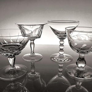 May include: Four clear crystal champagne coupe glasses with various etched and cut designs. The glasses have a classic stem and footed base, reflecting light. The coupe glasses are arranged on a reflective surface.