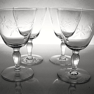 May include: Four clear wine glasses with starburst designs etched into the bowls. The glasses have rounded bowls, slender stems, and wide, flat bases. The glasses are arranged on a reflective surface.