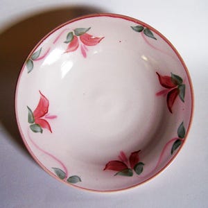 May include: A round ceramic bowl with a pale pink base color, featuring a hand-painted floral pattern of red petals and green leaves arranged around the interior rim. The bowl has a thin, solid red line along the outer edge of the rim.