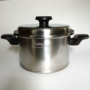 May include: A stainless steel cooking pot with a matching lid and two black side handles. The pot features the engraved text Lifetime Custom Designed Stainless Steel Made in U.S.A. under Patent on the front surface, with a black knob on the lid.