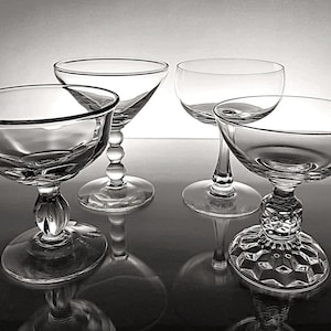 May include: Four clear champagne coupe glasses with varying stem and base designs. The glasses are arranged on a reflective surface, showcasing their elegant shapes and details. The stems include a beaded design, a smooth stem, and a faceted base.