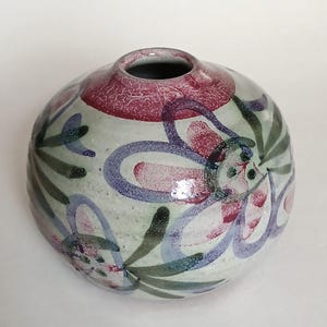 May include: A round ceramic vase with a light gray speckled base featuring hand-painted floral motifs in shades of purple, pink, and dark green. The top rim is finished with a textured, deep red circular band, creating a distinct contrast against the floral pattern that wraps around the spherical body of the vessel.