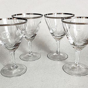 May include: A set of four clear glass cocktail glasses with a silver rim and an etched floral pattern. Each glass features a wide, V-shaped bowl, a slender stem, and a circular base. The delicate floral design is etched into the glass surface.