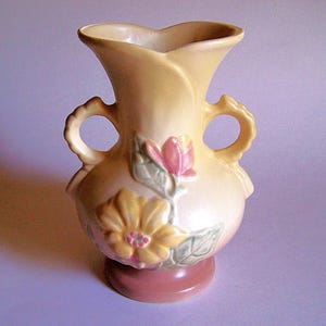 May include: A vintage ceramic vase in a soft yellow hue, featuring a flared top and two loop handles. The vase is adorned with embossed floral designs in pink and yellow, with green leaves. The base is a muted pink color, adding a touch of elegance to this decorative piece.