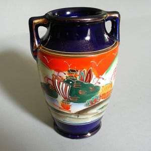 May include: A small ceramic vase with two handles, featuring a deep blue upper and lower section with gold trim. The central band displays a colorful landscape scene with green trees, red structures, and white birds against an orange sky.