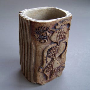 May include: A handmade, rectangular ceramic vase with a textured, bark-like exterior. The vase features carved relief designs of stylized faces and patterns. The interior is a lighter tan color, contrasting with the darker brown exterior.