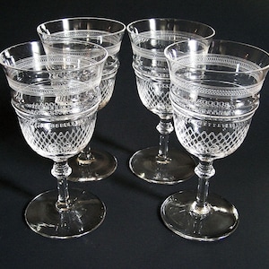 May include: Four clear, stemmed wine glasses with intricate etched designs. Each glass features a diamond pattern on the bowl and decorative bands around the rim and base. The glasses sit on a dark surface.