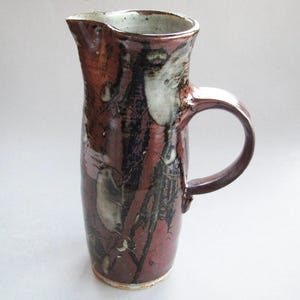 May include: A tall, handmade ceramic pitcher with a dark red and black mottled glaze. The pitcher has a large handle and a pouring spout. The design includes white and gray accents. A unique piece for serving beverages.