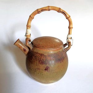May include: A ceramic teapot with a bamboo handle. The teapot is a rounded shape with a lid and a spout. The colors are earthy tones of brown, green, and red. The handle is attached with white wire.
