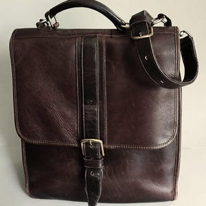 May include: A dark brown leather satchel featuring a top handle and an adjustable shoulder strap. The bag has a front flap secured by a vertical leather strap with a silver-toned metal buckle. The leather surface shows natural grain and wear.
