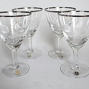 May include: A set of four clear glass stemmed goblets featuring a delicate etched floral pattern on the bowl and a polished silver rim. Each glass has a circular base with a small circular sticker attached to the center of the foot of the glass.