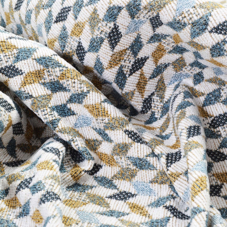 May include: Close-up of a white fabric with a geometric pattern in shades of blue and gold. The fabric is woven with a textured, slightly raised design.
